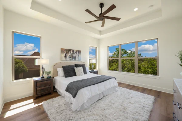 Primary bedroom with tray ceiling, wood ceiling fan, and windows on three sides overlooking green treetops