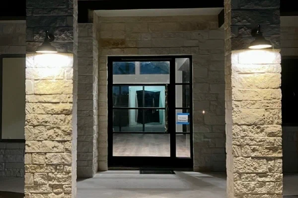 Limestone-clad entry of a Texas Hill Country custom home with black steel glass door and gooseneck sconces at night