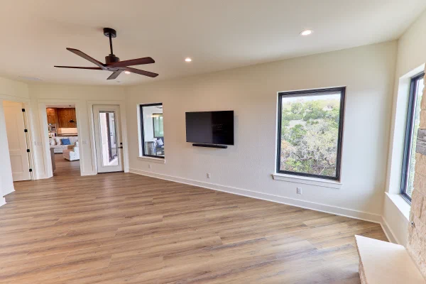 Media room with wall-mounted TV between black-framed picture windows overlooking Texas Hill Country greenery