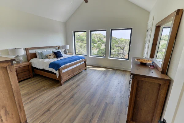 Primary bedroom with vaulted ceiling and three black-framed windows overlooking Texas Hill Country treetops