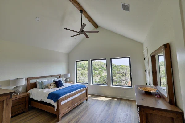 Vaulted primary suite with exposed wood beam, dark wood ceiling fan, and wall of windows to wooded hills