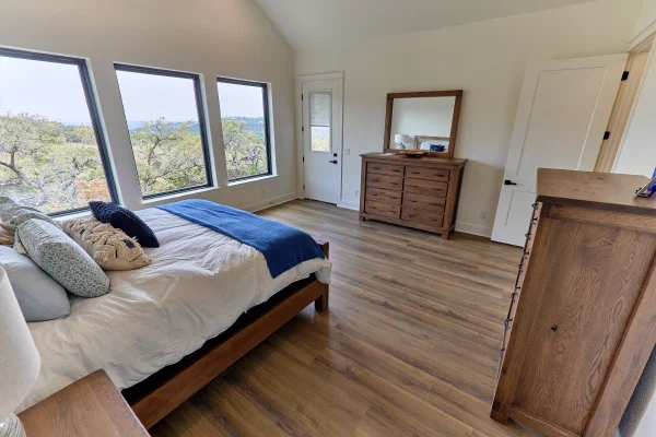 King bed under a trio of black-framed windows with wooded views, oak dresser with mirror, and patio door