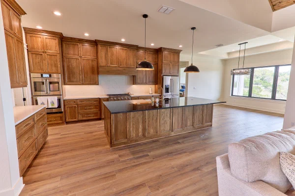 Warm oak shaker cabinets surround a black granite island under bronze dome pendants with tray-ceiling dining beyond