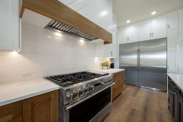 Stainless gas range under a wood-trimmed vent hood with glossy white subway tile backsplash and built-in refrigerator