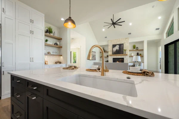 Gold gooseneck faucet over an undermount sink in veined quartz with dark island cabinets and a cozy fireplace beyond