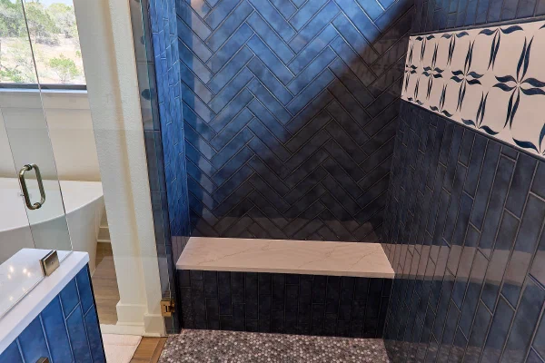 Walk-in shower with blue herringbone walls, patterned accent band, quartz seat, and gray penny round tile floor