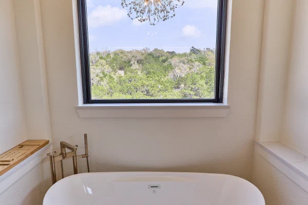 Freestanding soaking tub centered under a window, with crystal chandelier, brass floor-mount filler, and bath caddy