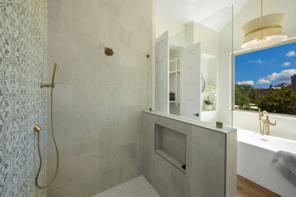 Walk-in shower with brass fixtures, patterned accent tile, stone-look walls, and glass partition beside a soaking tub